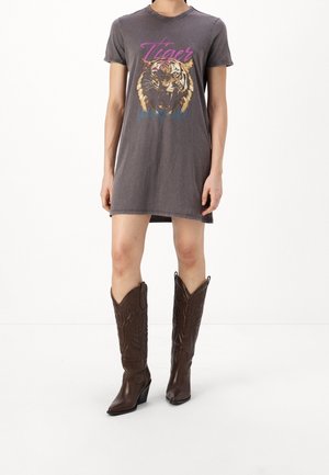 Kvinde iført en falmet mørkegrå t-shirt-kjole med tigergrafik og tekst, kombineret med knæhøje brune cowboystøvler.