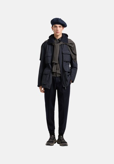 Giacca imbottita blu navy con multiple tasche, felpa grigia con cappuccio, pantaloni scuri e sneakers nere. Il modello indossa un berretto blu navy. Look casual a strati.