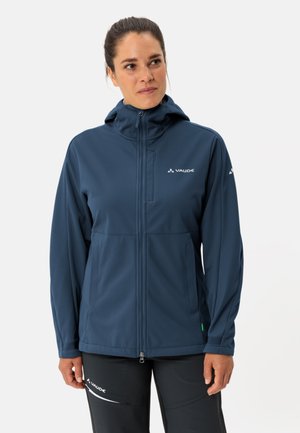 Femme portant une veste à capuche zippée bleu marine et un pantalon noir avec des logos Vaude, debout devant un fond clair.