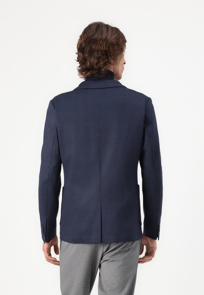Homme avec des cheveux ondulés brun moyen portant un blazer bleu marine ajusté et un pantalon gris clair, se tenant de dos contre un fond blanc uni.