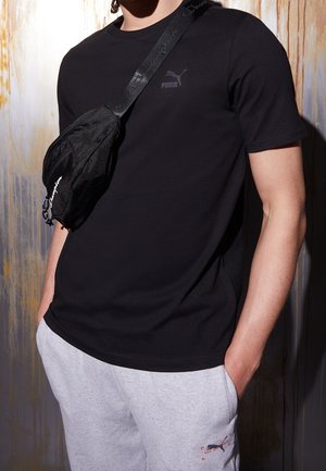 T-shirt nera a maniche corte con un sottile logo Puma e pantaloni della tuta grigi con stampa decorativa. Il modello porta una borsa a tracolla nera.