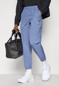 Αθλητικά παντελόνια σε ανοιχτό μπλε χρώμα με ελαστική μέση, πλαϊνές τσέπες και ελαστικά μανίκια; συνδυασμένα με μια μαύρη τσάντα και λευκά αθλητικά παπούτσια.