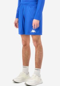 Blå idrottshorts med elastisk midjeband och vit logotyp; kombinerade med vita sneakers och mönstrade strumpor. Slätt, lättviktigt tyg.