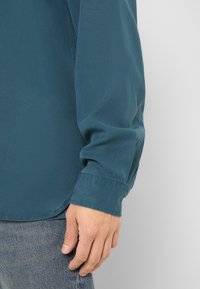 Main masculine reposant à côté du poignet d'une chemise bleue à manches longues et d'un jean en denim bleu clair sur un fond uni.