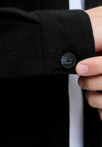Fondo di una camicia nera in lana, con un polsino che presenta un bottone nero rotondo a due fori, tenuto tra le dita e che mostra una superficie di tessuto testurizzato.