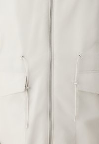 Lys beige jakke med hel lynlås, to frontlommer og justerbare snorer i kanten. Glatt, vannavstøtende stoff.