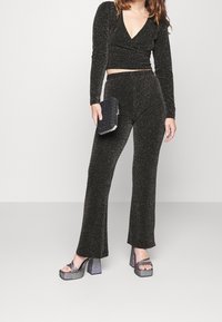 Svart, glittrande tvådelad outfit med en djup V-ringad långärmad kort topp och utsvängda byxor. Kompletteras av en svart kuvertväska och metalliska plattformsskor.