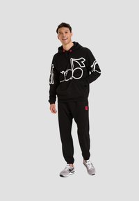 Felpa nera con design grafico bianco, joggers neri e sneakers grigie. Presenta cappuccio, coulisse e un accento rosso sul logo dei pantaloni.