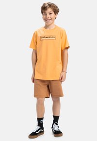 Oranžové bavlnené tričko s obdélníkovým logom, doplnené hnedými šortkami. Čiernobiele tenisky a čierne ponožky dotvárajú tento outfit.
