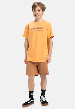 T-shirt en coton orange avec un logo rectangulaire, associé à un short marron. Des chaussures noires et blanches et des chaussettes noires complètent la tenue.