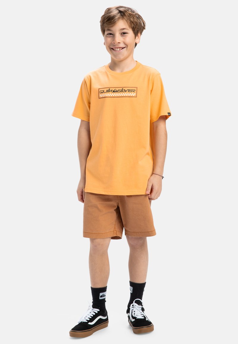Oranžové bavlnené tričko s obdélníkovým logom, doplnené hnedými šortkami. Čiernobiele tenisky a čierne ponožky dotvárajú tento outfit.