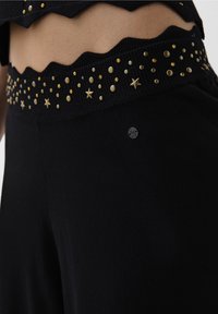 Sorte bukser med en ribbet talje, prydet med guldfarvede studs og stjerneaccenter. Materialet ser blødt ud med et afslappet design.