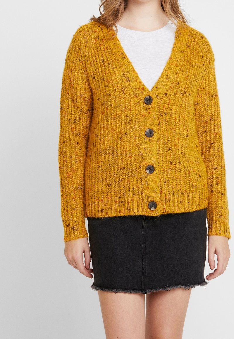 Cardigan a maglia giallo senape con scollo a V, cinque bottoni scuri, texture a coste e maniche lunghe, abbinato a una gonna in denim nera, strappata.