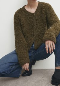 Olijfgroene, donzige cardigan met ronde hals, gouden knopen en oversized mouwen. Gecombineerd met blauwe spijkerbroek en zwarte enkellaarsjes.