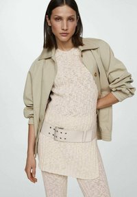 Kvinna med rakt mörkt hår bär en beige stickad topp och byxor med breda bälten samt en ljusbeige jacka, poserande med handen på höften.