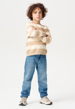 Beige und weiße gestreifte Pullover, weicher Stoff, Rundhalsausschnitt. Hellblaue Jeans. Beige und graue gemusterte Turnschuhe. Kind steht selbstbewusst da.
