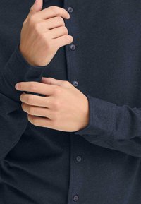 Mørk marineblå knappskjorte laget av mykt stoff, med en tettsittende design som har knapper langs fronten og veldefinerte mansjetter.