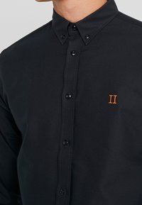 Sort skjorte med knapper lavet af glat stof, med en klassisk krave, sorte knapper og et orange broderet "II" på brystet.