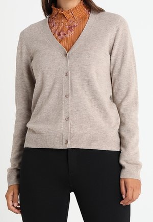 Femme portant un cardigan beige boutonné à col en V sur une blouse orange texturée à col haut, associée à un pantalon noir.