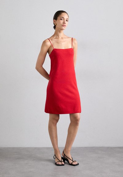 Rode mouwloze jurk met een vierkante halslijn, eenvoudige silhouet en een licht uitlopende zoom. Model draagt zwarte sandaaltjes met straps.