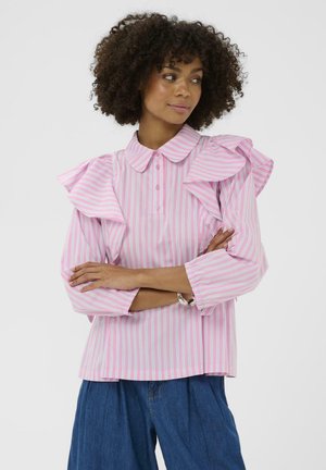 Femme aux cheveux bouclés portant un chemisier rayé rose et blanc avec des épaules à volants et un pantalon bleu à jambes larges, bras croisés, regardant vers la droite.