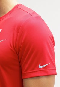 Czerwona koszulka sportowa wykonana z oddychającego materiału o teksturowanej powierzchni. Wyposażona w srebrne logo Nike na lewym rękawie. Krótkie rękawy i okrągły dekolt.