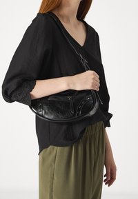 Borsa a spalla in pelle nera con forma curvata, tasca con zip e dettagli in metallo. Abbinata a una blouse nera e pantaloni verde oliva.
