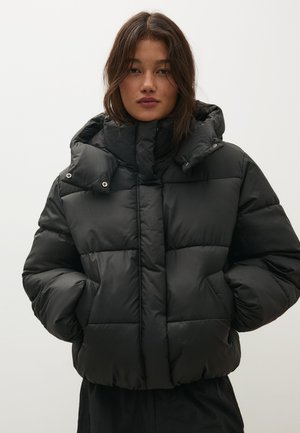 Frau mit mittellangem dunklem Haar trägt eine schwarze Steppjacke mit hohem Kragen und Händen in den Taschen vor einem einfarbigen hellen Hintergrund.