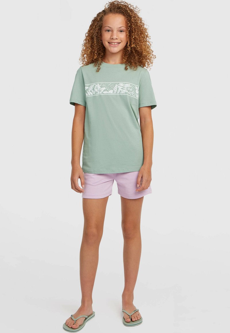 Hellgrünes T-Shirt mit kurzen Ärmeln und weißem Blumenmuster, kombiniert mit lila Shorts und grünen Flip-Flops. Glatte Stoffstruktur.