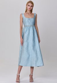 Robe bleu clair, longueur genoux, avec une surface texturée, décolleté carré, larges bretelles, et une taille ornée de perles, assortie de sandales blanches.