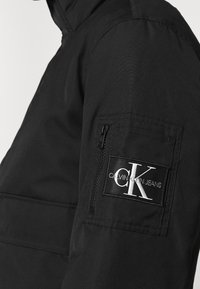 Svart ytterjacka med en texturerad yta, försåld med en ficka med dragkedja på ärmen och en logotypapplikation som visar "Calvin Klein Jeans."