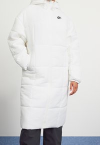 Vit vadderad lång kappa med dragkedja, huva och svart logotyp på bröstet. Har elastiska ärmslut och en quiltad textur.