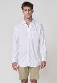 Cămașă albă cu mânecă lungă, cu nasturi, dotată cu un buzunar pe piept, confecționată dintr-un material ușor. Asortată cu pantaloni scurți khaki. Design curat și simplu.
