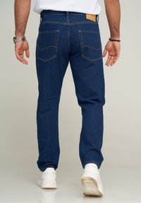 Man som bär mörkblå jeans, vita sneakers, silverur på vänster handled och svart pärlarmband på höger handled, står på ett ljust golv.