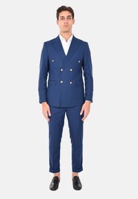 Abito blu doppiopetto con bottoni dorati, tessuto testurizzato, vestibilità slim e pantaloni con risvolto, abbinato a una camicia bianca e scarpe nere.