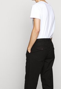 Osoba v bílé košili s krátkým rukávem, zasunuté do černých kalhot s vysokým pasem a zapínacími zadními kapsami, stojící s rukama v kapsách.