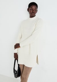 Pull en maille côtelée couleur crème avec col montant et fentes sur les côtés, associé à un pantalon beige et un petit sac à main noir.