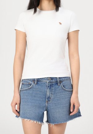 T-shirt blanc à manches courtes avec un petit logo brodé marron, associé à un short en denim bleu avec un ourlet effiloché et des poches avant.