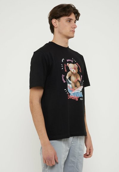 Young man in black t-shirt with colorful teddy bear surfing graphic, standing against plain light background, facing right.