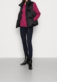 Chaleco acolchado negro, camiseta de manga larga de color burdeos, jeans ajustados oscuros y botas de tacón negras. Cuenta con un cuello alto y cierre de cremallera.