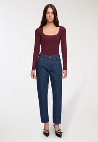 Kobieta stoi w dopasowanej bordowej bluzce z długim rękawem, niebieskich jeansach z wysokim stanem i czarnych szpilkach na białym tle.