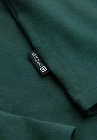 Dunkelgrünes Baumwoll-T-Shirt mit einer glatten Textur. Verfügt über einen runden Halsausschnitt und ein kleines schwarzes Etikett mit dem Markennamen "ombre", das an der Seite genäht ist.