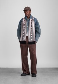 Džinsi jaka ar platām piedurknēm, balta krekls, brūni uzpildes bikses, melni apavi un rozā-melns frinģu šalle ar tekstu "HANK KØBENHAVN".