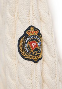 Kabelstickad krämfärgad tröja med en rund, broderad patch med en krona, "POLO CLUB", och dekorativa element i blått, rött och guld.