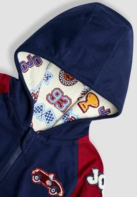 Hanorac cu fermoar, cu corp navy și mâneci roșii, dotat cu o pată cu mașină, brodat "JOJO" și interior cu un model ce prezintă motive de curse.