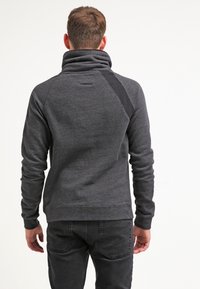 Homme portant un pull à col haut gris foncé avec un panneau noir sur l'épaule, associé à un jean noir, vu de dos sur un fond blanc.
