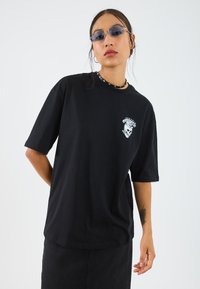 T-shirt en coton noir à manches courtes, avec un motif de skull blanc sur le côté gauche de la poitrine. Porté avec une jupe en denim noir et des accessoires.