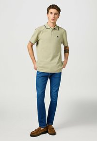 Olivgrön polo-shirt med svart och vit randig krage, kort ärm och logotyp. Matchad med blå jeans och bruna mockaskor.