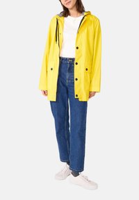 Veste de pluie imperméable jaune avec capuche, ornée de boutons noirs et de deux poches avant, portée sur une chemise blanche et un jean bleu.