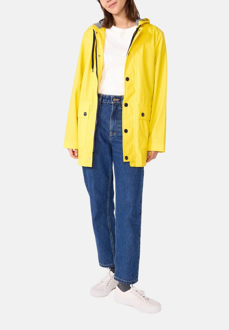 Veste de pluie imperméable jaune avec capuche, ornée de boutons noirs et de deux poches avant, portée sur une chemise blanche et un jean bleu.